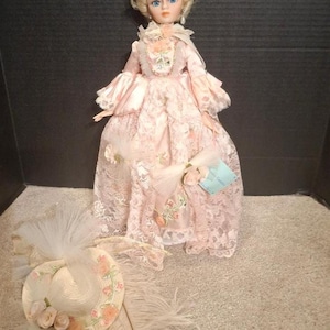 May include: A porcelain doll with blonde hair and blue eyes wearing a pink satin gown with lace overlays and floral accents, accompanied by a matching wide-brimmed hat decorated with fabric roses, tulle, and a long white feather plume.