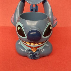 May include: Blue Stitch character shaped plastic cup with a removable lid. The cup has a red collar with a gold emblem that says "Ohana".