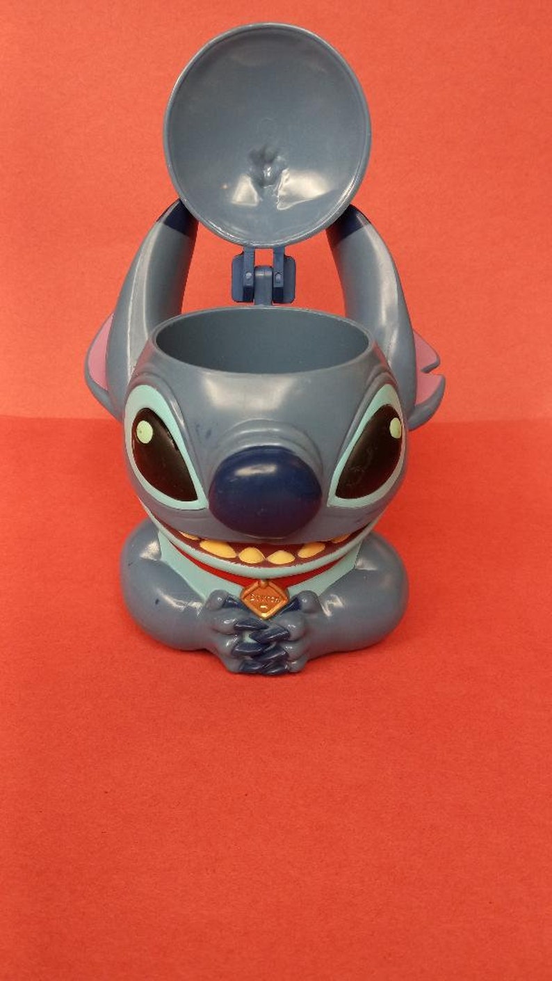 May include: Blue Stitch character shaped plastic cup with a removable lid. The cup has a red collar with a gold emblem that says "Ohana".