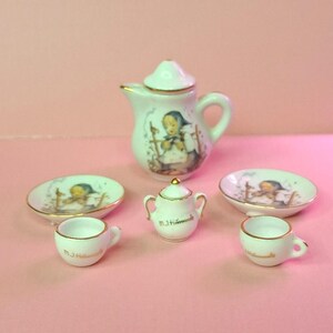 May include: A miniature porcelain tea set with a white base and gold trim. The set includes a teapot, sugar bowl, two saucers, and two teacups. Each piece features a painted image of a girl and the text "M.J. Hummel".