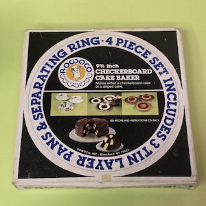 May include: A square cardboard box for a Rowoco 9 3/4 inch checkerboard cake baker. The black and white packaging features blue text, a logo of a chef, and illustrations of metal cake pans and a finished chocolate cake with a checkerboard pattern.