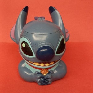 May include: Blue Stitch character figurine with a red collar and a gold charm. The character has large eyes, a wide smile, and a blue nose.