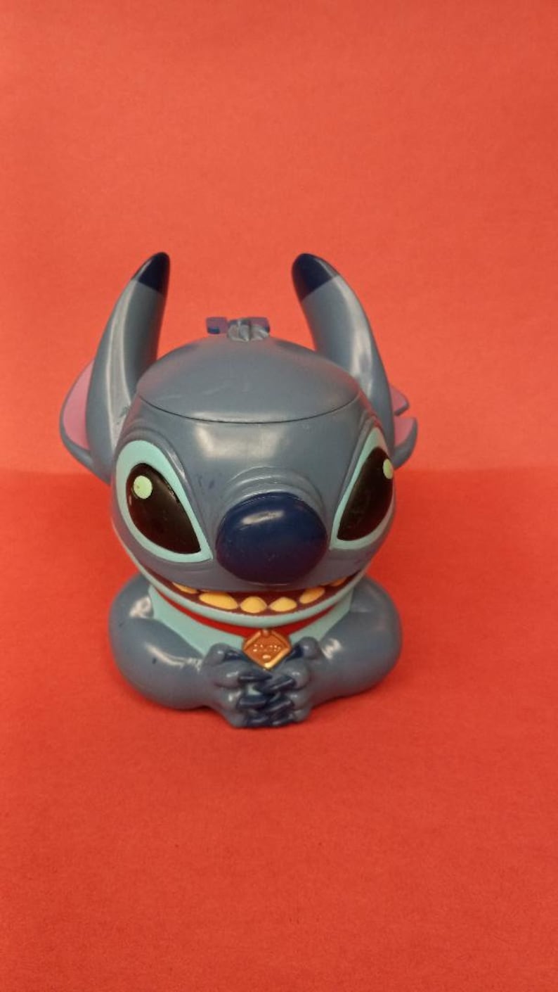 May include: Blue Stitch character figurine with a red collar and a gold charm. The character has large eyes, a wide smile, and a blue nose.