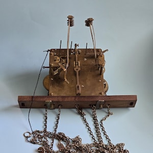 May include: A brass clock movement mechanism featuring an exposed gear train, two vertical hammer arms, and a dark wooden mounting plate. Three long metal chains hang from the base, each equipped with small metal weights and hooks for suspension.