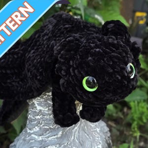 Könnte beinhalten: Ein schwarzes gehäkeltes Plüschtier mit leuchtend grünen Augen. Das Spielzeug hat die Form eines Drachen oder einer Katze. Das Wort "PATTERN" ist auf einem blauen Banner oben links aufgedruckt.