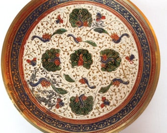 May include: A round brass plate with a white background and intricate floral and peacock designs in shades of green, blue, red, and gold. The plate has a raised rim with a blue and gold border.