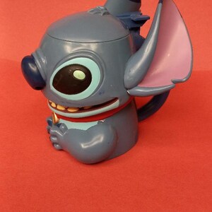May include: Blue Stitch character mug with a red collar and a gold tooth. The mug has a blue and purple ear and a blue lid.