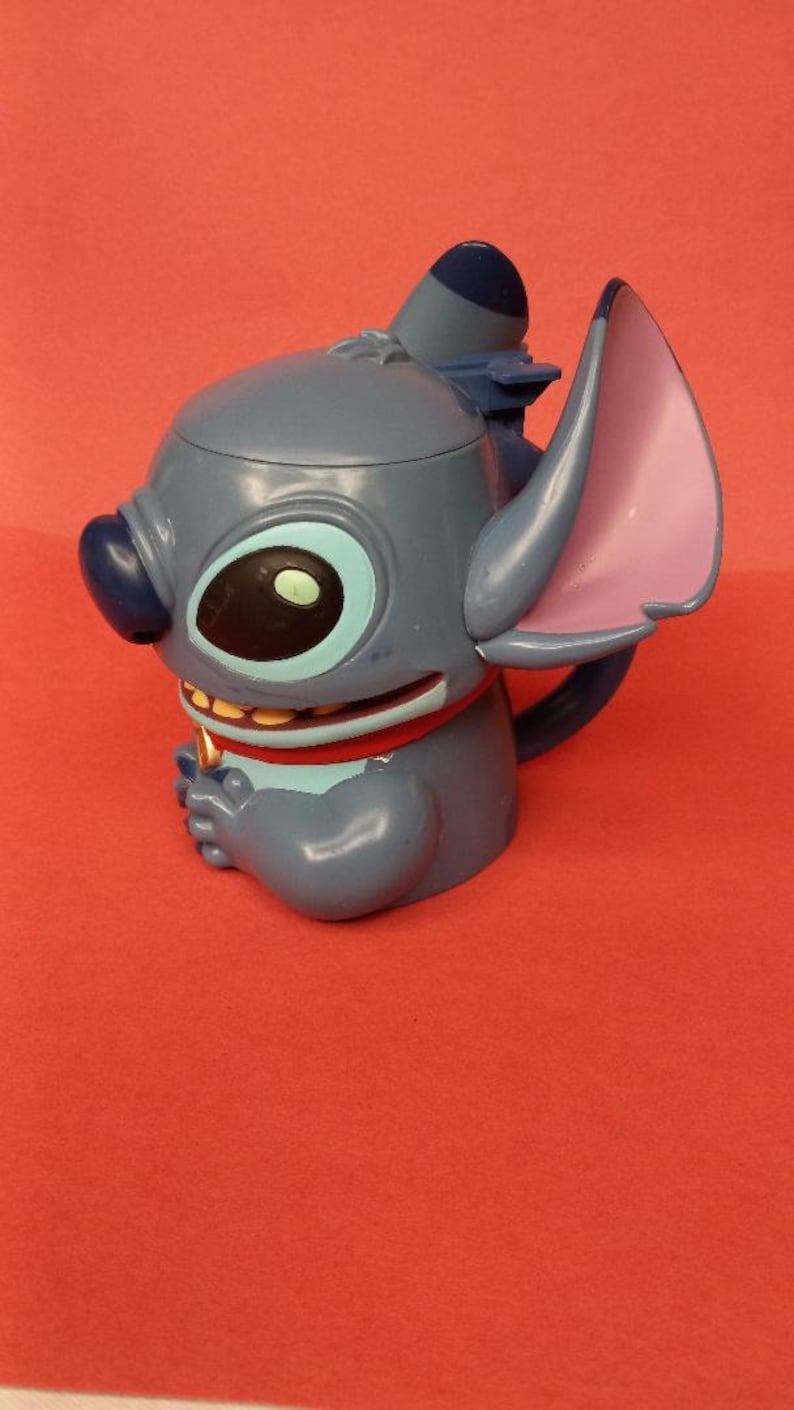 May include: Blue Stitch character mug with a red collar and a gold tooth. The mug has a blue and purple ear and a blue lid.