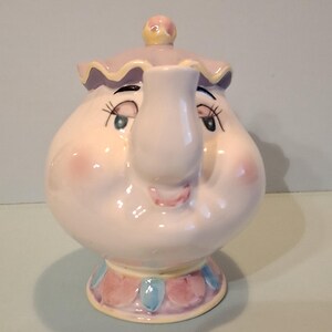 May include: A ceramic teapot shaped like a character with a round, pale pink body, a spout, and a lid. The teapot features painted facial details including closed eyes with long eyelashes, rosy cheeks, and a small mouth. The base is decorated with pink and blue petal-like shapes, and the lid has a ruffled edge with a small yellow knob.