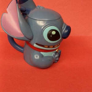 May include: Blue Stitch character mug with a pink ear and a red collar. The mug is made of plastic and has a handle.