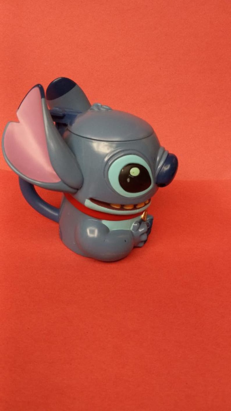 May include: Blue Stitch character mug with a pink ear and a red collar. The mug is made of plastic and has a handle.