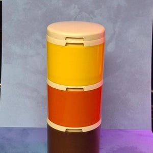 May include: A stackable storage container with three sections in yellow, orange, and brown. Each section has a white band and a cream-colored lid. The container is cylindrical and sits on a blue and purple background.