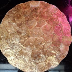 May include: A round, decorative plate with a textured surface. The plate features a pattern of overlapping, shell-like shapes in shades of gold and pink. The edges of the plate are scalloped.