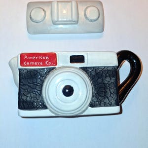 May include: A white ceramic teapot shaped like a vintage camera with black detailing and a red label that reads "American camera co.". The teapot has a spout and a handle. The lid is separate and is also white ceramic with two round knobs.