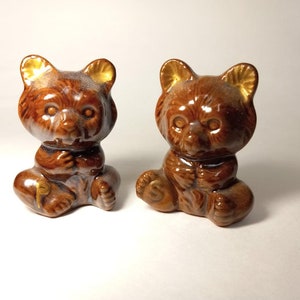 May include: Two brown ceramic bear figurines with golden accents on their ears. The bears are sitting with their paws clasped, and have a glossy finish. These decorative figurines are ideal for a shelf or display.
