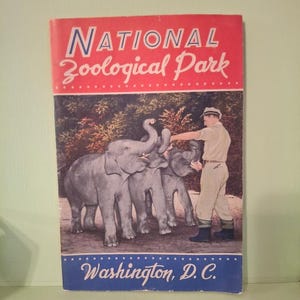 May include: A rectangular brochure titled National Zoological Park Washington, D.C. The cover features a color illustration of a zookeeper in khaki clothing feeding three gray elephants in a wooded area with a red top and blue bottom border.