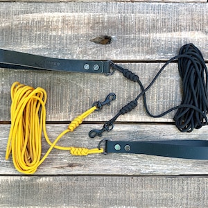 Puede incluir: Dos correas de cuero negro para perros con clips metálicos y una correa de cuerda amarilla con una correa de cuerda negra unida a ella. Las correas están enrolladas en una superficie de madera.