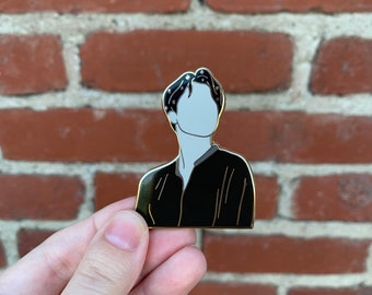 May include: A black enamel pin featuring a stylized illustration of a person's upper body. The figure has a light gray face and dark hair with small star accents. The pin has a gold border and is set against a brick background.