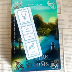 May include: A bookmark with light blue postage stamp designs featuring a deer, stars, and a dragon, rests on a book titled "Empire of Storms" by Sarah J. Maas. The bookmark has text that reads "welcome to terrasen", "you could rattle the stars", and "the thirteen".