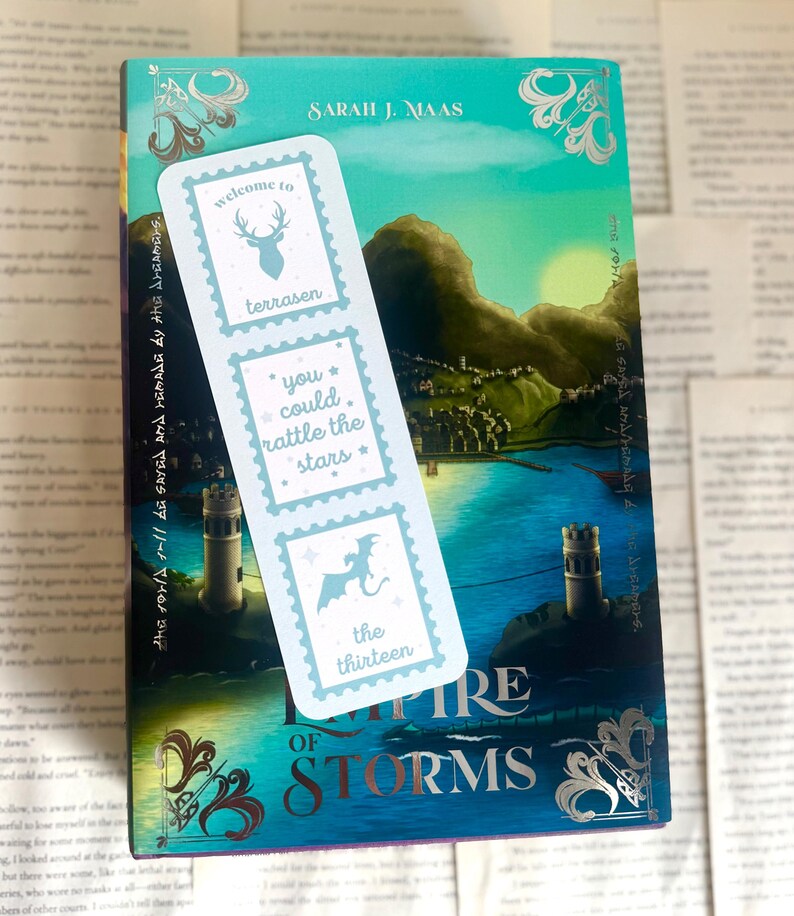 May include: A bookmark with light blue postage stamp designs featuring a deer, stars, and a dragon, rests on a book titled "Empire of Storms" by Sarah J. Maas. The bookmark has text that reads "welcome to terrasen", "you could rattle the stars", and "the thirteen".