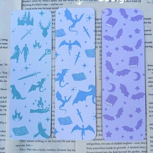 May include: Three rectangular bookmarks with double-sided designs. One is light blue with castle, sword, and dragon motifs. Another is light blue with dragon, book, and sword patterns. The third is lavender with bats and book designs.