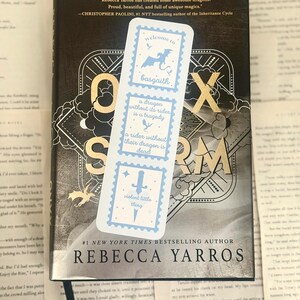 May include: A bookmark with a light blue design featuring a dragon, sword, and text from the book "Iron Flame" by Rebecca Yarros. The bookmark is placed on top of the book. The book cover is dark with gold lettering.