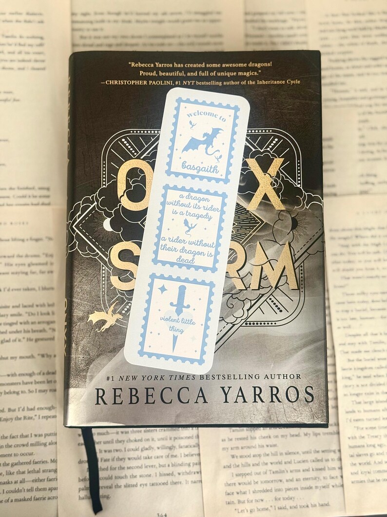 May include: A bookmark with a light blue design featuring a dragon, sword, and text from the book "Iron Flame" by Rebecca Yarros. The bookmark is placed on top of the book. The book cover is dark with gold lettering.