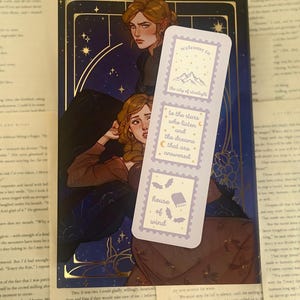 May include: A bookmark with a light purple background and text that reads "welcome to the city of starlight", "to the stars who listen and the dreams that are answered", and "house of wind". The bookmark is placed on top of a book with a dark blue cover and illustrations.