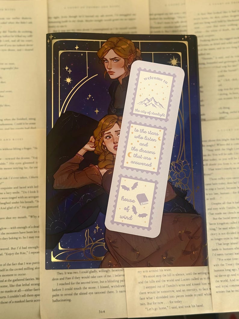 May include: A bookmark with a light purple background and text that reads "welcome to the city of starlight", "to the stars who listen and the dreams that are answered", and "house of wind". The bookmark is placed on top of a book with a dark blue cover and illustrations.