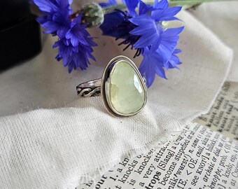 May include: A silver ring featuring a smooth, pale green, teardrop-shaped stone set in a bezel. The band has a decorative twisted metal pattern. The ring is positioned on a piece of paper with printed dictionary text and blue cornflowers.