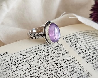 May include: A silver ring featuring an oval-shaped, light purple cabochon gemstone. The metal band is decorated with an intricate, embossed floral pattern. The ring is resting on an open dictionary page with visible text entries.