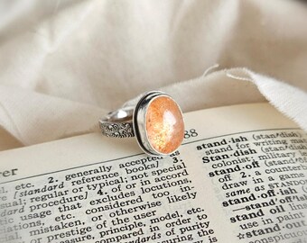 May include: A silver ring featuring an oval-shaped, speckled orange sunstone cabochon set in a smooth bezel. The ring band has a textured, engraved pattern along its outer surface, providing a detailed contrast to the polished stone.