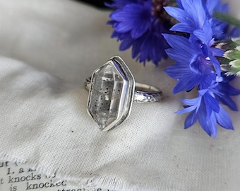May include: A silver ring featuring a hexagonal clear crystal with dark inclusions, set in a metal bezel on a textured band. The ring is positioned next to two vibrant blue cornflowers on a piece of white fabric resting on an open dictionary.