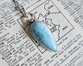 Puede incluir: Un colgante de piedra Larimar azul claro en forma de lágrima engastado en un marco de metal plateado. El colgante cuelga de una fina cadena plateada y descansa sobre una página de diccionario abierta con un mapa de la región de Khyber Pass.
