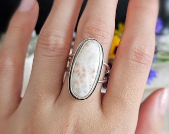 May include: A silver ring featuring a large, oval-shaped, cream-colored stone with subtle brown speckles. The stone is set in a smooth silver bezel, and the band consists of two thin, parallel silver wires that meet at the sides of the setting.