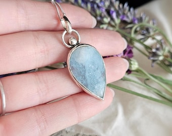 May include: A teardrop-shaped pendant featuring a pale blue, semi-translucent stone set in a polished silver frame. The pendant hangs from a delicate silver chain and includes a small silver bead accent at the top where the chain connects.