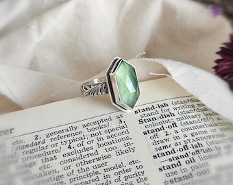 May include: A silver ring featuring a faceted, hexagonal light green stone set in a bezel. The band of the ring displays a detailed, embossed leaf-like pattern along its outer surface, resting on an open book page with visible printed text.