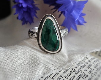May include: A silver ring featuring a teardrop-shaped malachite gemstone with dark green banded patterns. The metal band displays an intricate engraved leaf or vine motif along the sides, providing a detailed texture to the polished silver setting.