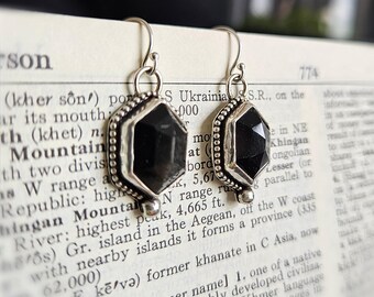 Puede incluir: Un par de pendientes colgantes de plata con piedras negras hexagonales facetadas engastadas en un marco de metal con cuentas. Los pendientes cuelgan de ganchos de plata simples sobre una página de diccionario impresa con texto visible.