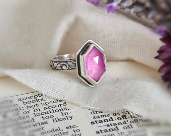May include: A silver ring featuring a faceted, hexagonal pink gemstone set in a metal bezel. The ring band is crafted from silver with an intricate, embossed floral pattern along the outer surface, providing a detailed texture to the metal.