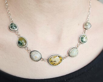 May include: A silver chain necklace featuring seven bezel-set, multi-colored gemstones in various shapes and sizes. The stones display earthy tones of green, yellow, and cream with natural inclusions, connected by silver links in a symmetrical arrangement.