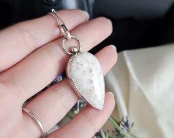 May include: A teardrop-shaped pendant featuring a smooth, off-white stone with a subtle, mottled texture, encased in a polished silver setting. The pendant hangs from a delicate silver chain, connected by a circular silver bail and a small bead.