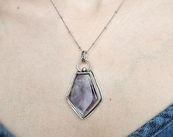 May include: A silver necklace featuring a pentagonal pendant with a polished purple and white stone set in a metal frame. The pendant hangs from a delicate silver chain with small evenly spaced metal beads along the length of the chain.