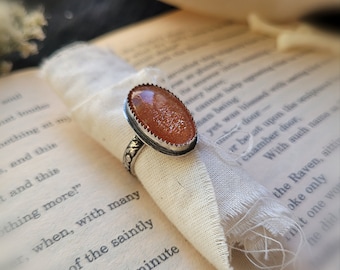 Puede incluir: Un anillo de plata con una piedra de sol naranja de forma ovalada engastada en un engaste de plata. El anillo está sobre un fondo de tela blanca.
