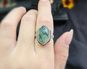 May include: A silver ring featuring a large, hexagonal green stone with intricate, natural patterns. The stone is set in a silver bezel with decorative detailing on the band. The ring's design showcases a unique, organic aesthetic.