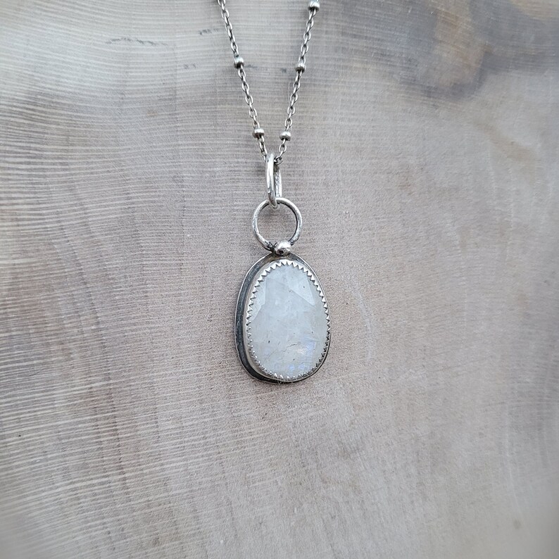 Puede incluir: Un collar de cadena de plata con un colgante que presenta una piedra de luna blanca engastada en un bisel de plata. La piedra de luna tiene un sutil brillo iridiscente.