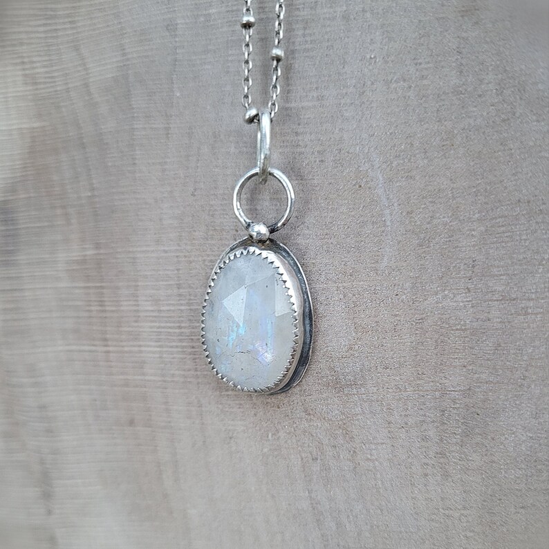 Puede incluir: Un collar de cadena de plata con un colgante de piedra lunar blanca. El colgante tiene forma ovalada y tiene un engaste de bisel de plata.