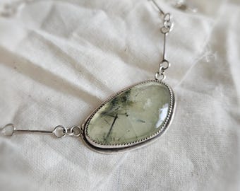 May include: A necklace featuring a polished, asymmetrical, pale green gemstone with dark needle-like inclusions, set in a serrated silver bezel. The pendant is attached to a delicate silver chain with thin, straight metal bar links.