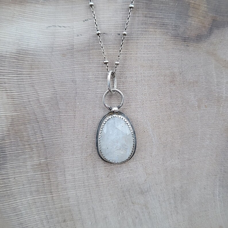 Puede incluir: Un collar de cadena de plata con un colgante ovalado blanco con un cristal transparente. El colgante est&aacute; engastado en un bisel de plata y tiene una delicada argolla de plata.