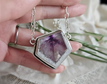 May include: A pentagonal pendant necklace featuring a faceted purple and white amethyst stone set in a polished silver metal frame. The pendant is suspended from a delicate silver chain with oval links, showcasing a natural crystalline texture.